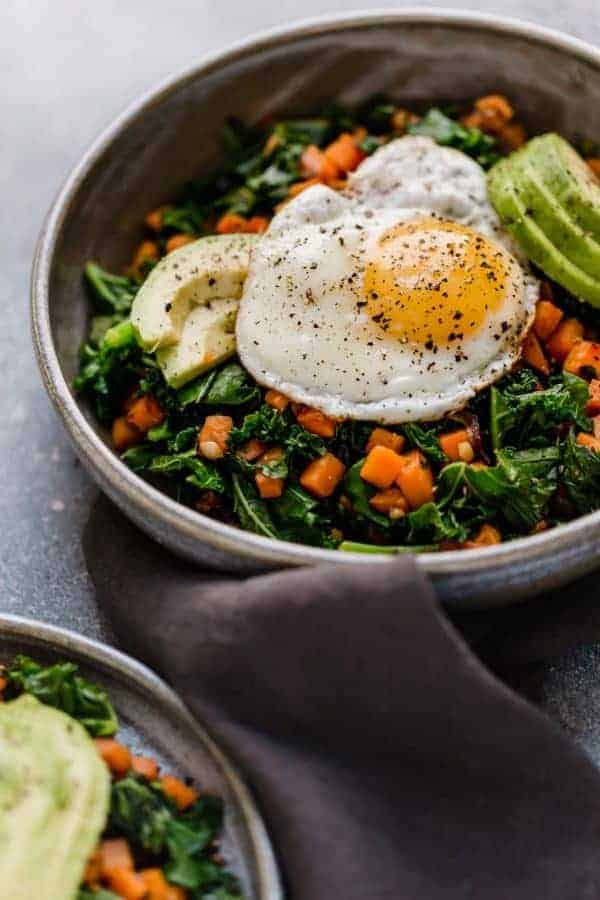 Photo take from an angle of the Kale and Sweet Potato Sauté in a bowl with a fried egg on top and avocado slices.
