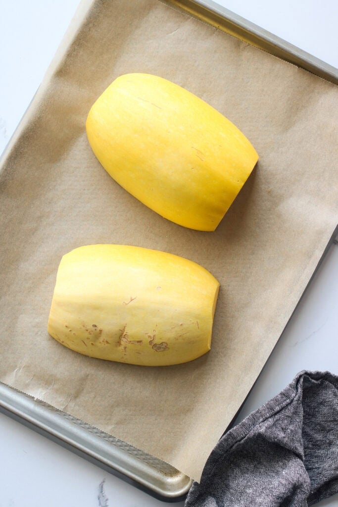 Overhead view of a baking sheet lined with parchment paper and two halves of a spaghetti squash face down ready for baking. 