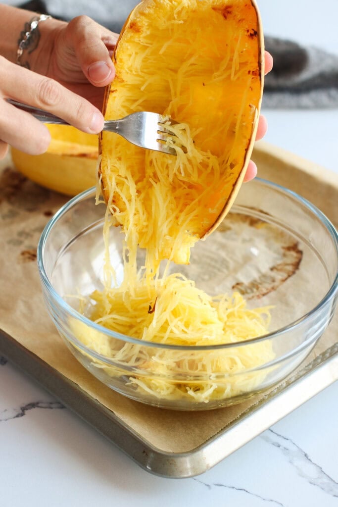 Close up view of hands holding and scooping out freshly baked spaghetti squash into a glass bowl. 