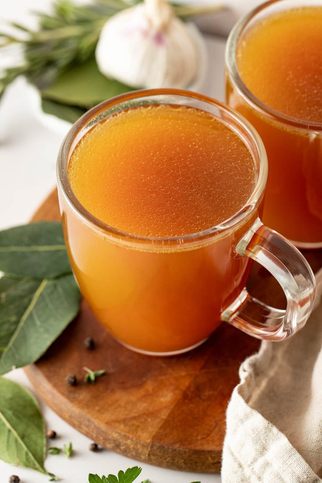 Close up view of a glass mug filled with a rich bone broth next to bay leaves on a wooden cutting board.