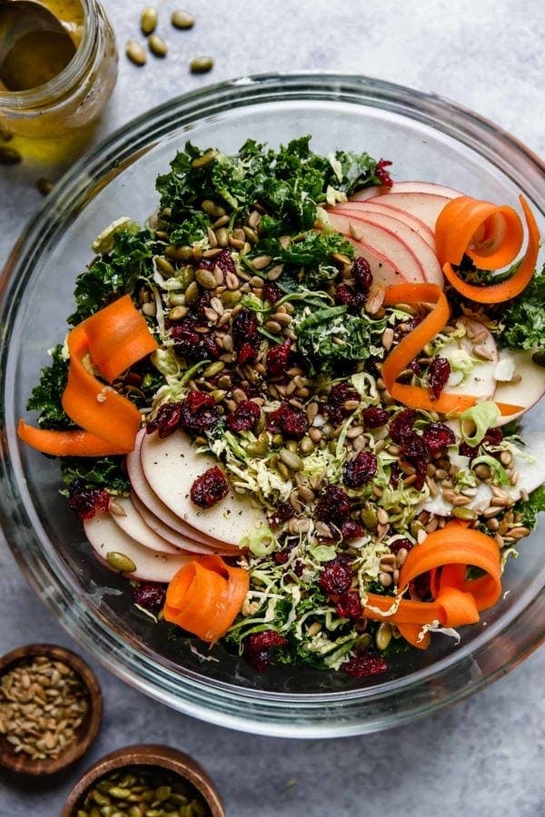 Overhead view of a glass bowl filled with Massaged Kale Salad topped with sliced apples, carrots, and dried cranberries.