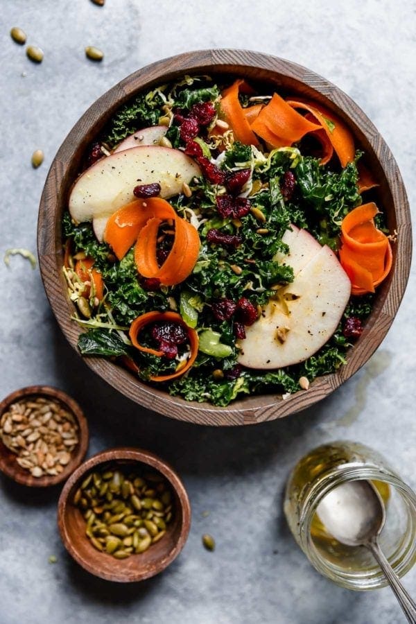 Overhead view of a wooden bowl of Massaged Kale Salad topped with sliced carrots, apples, and dried cranberries. 