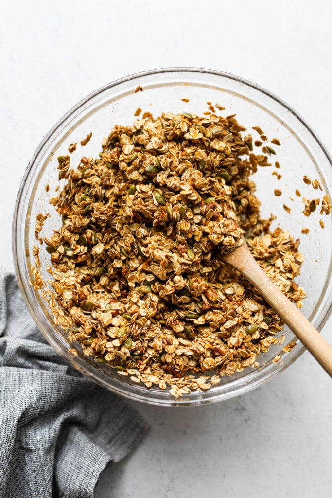 Pumpkin granola mixed together in a clear glass mixing bowl with a wooden spoon