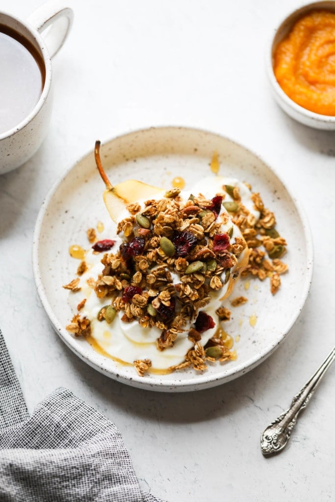 Pumpkin spice granola with dried cranberries and pecans over a yogurt topped baked pear half on a speckled plate. 