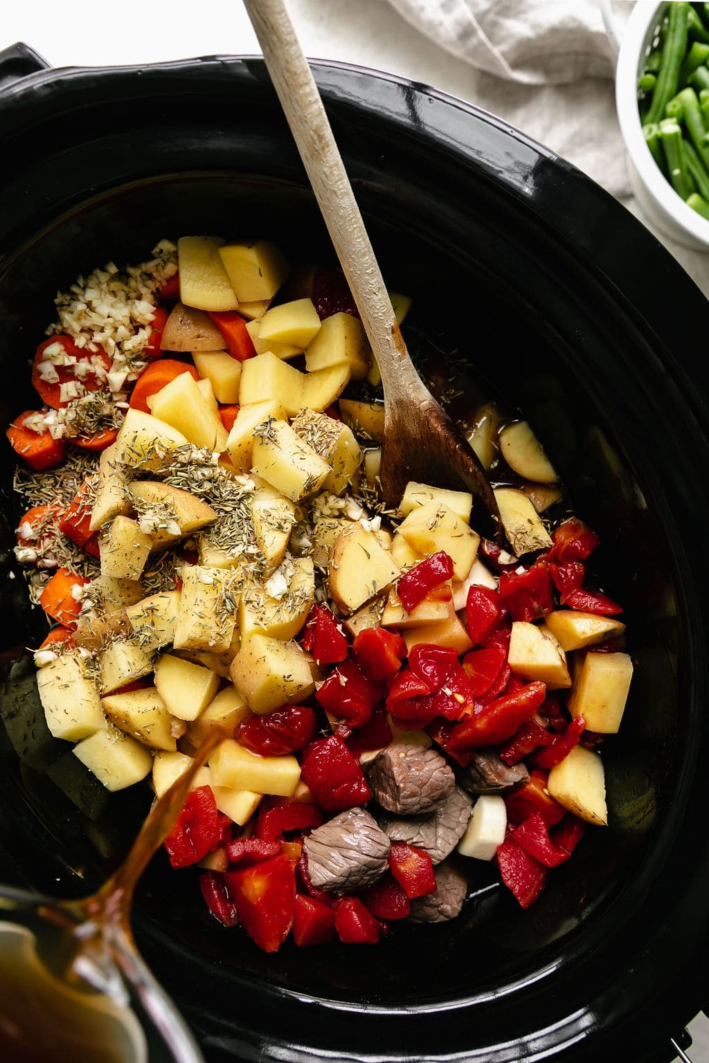 All ingredients for slow cooker beef stew with root vegetables in black slow cooker, broth being poured in
