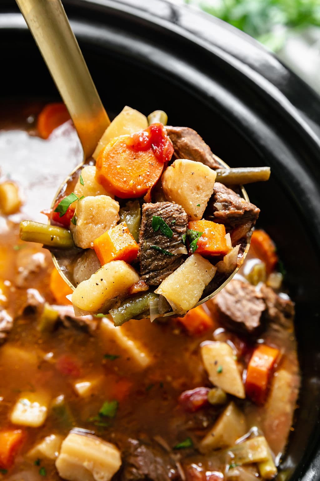 A ladle full of beef stew with potatoes, parsnips, and green beans over a black slow cooker. 