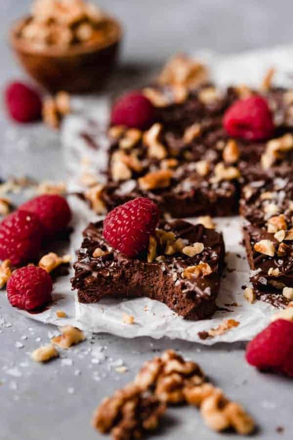 Photo of the Vegan No-Bake Brownies with one having a perfect bite out of it, on a piece of white parchment with raspberries, walnuts and chocolate drizzle.