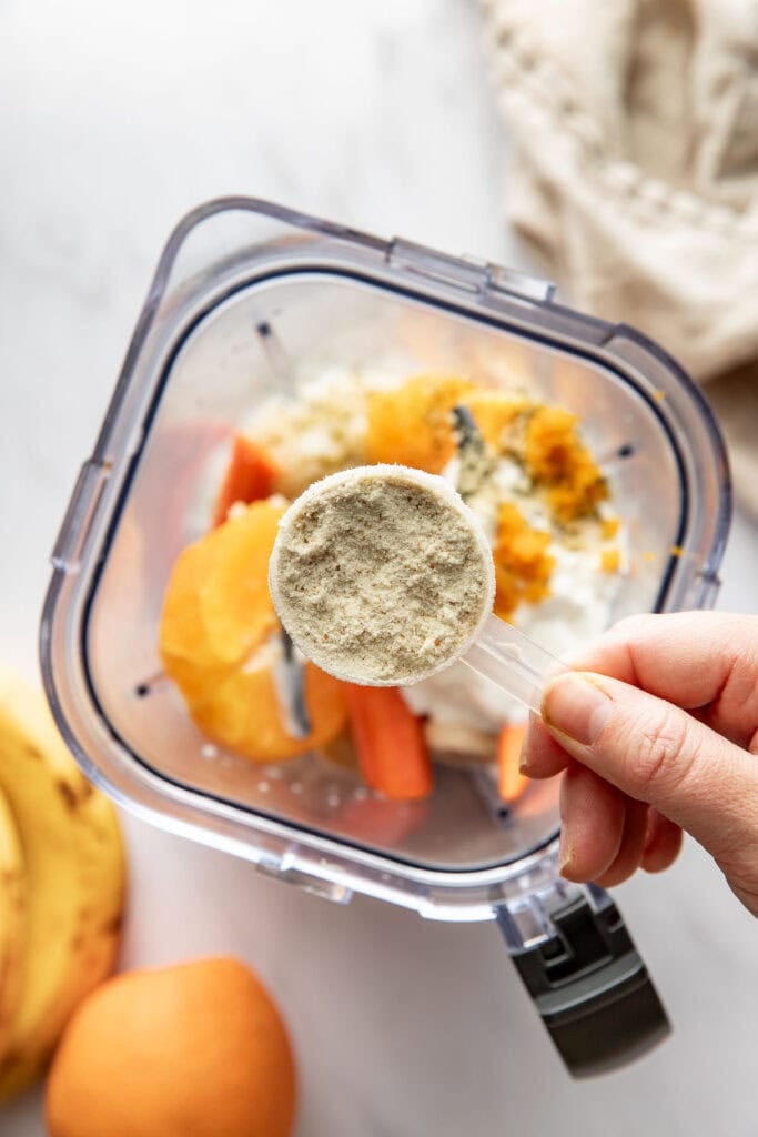 Overhead view of the inside of a blender filled with orange smoothie ingredients including a scoop of protein powder.