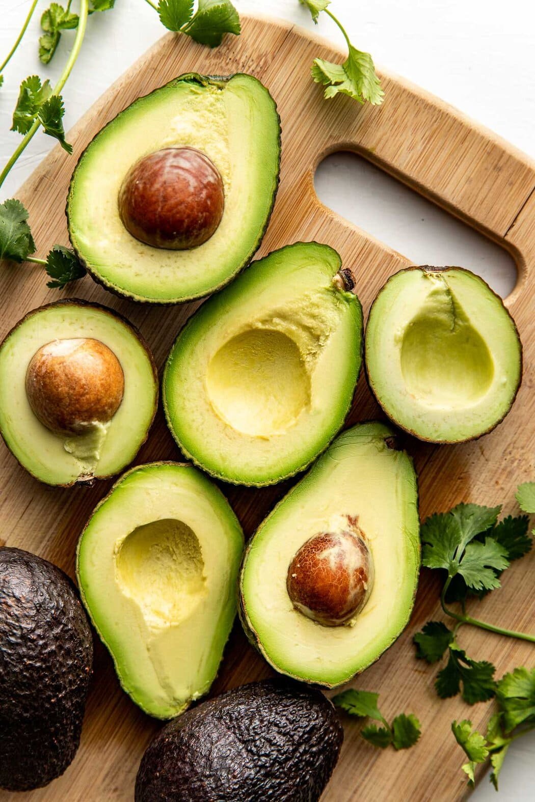 Overhead view of halved avocados on a wooden cutting board surrounded by fresh cilantro