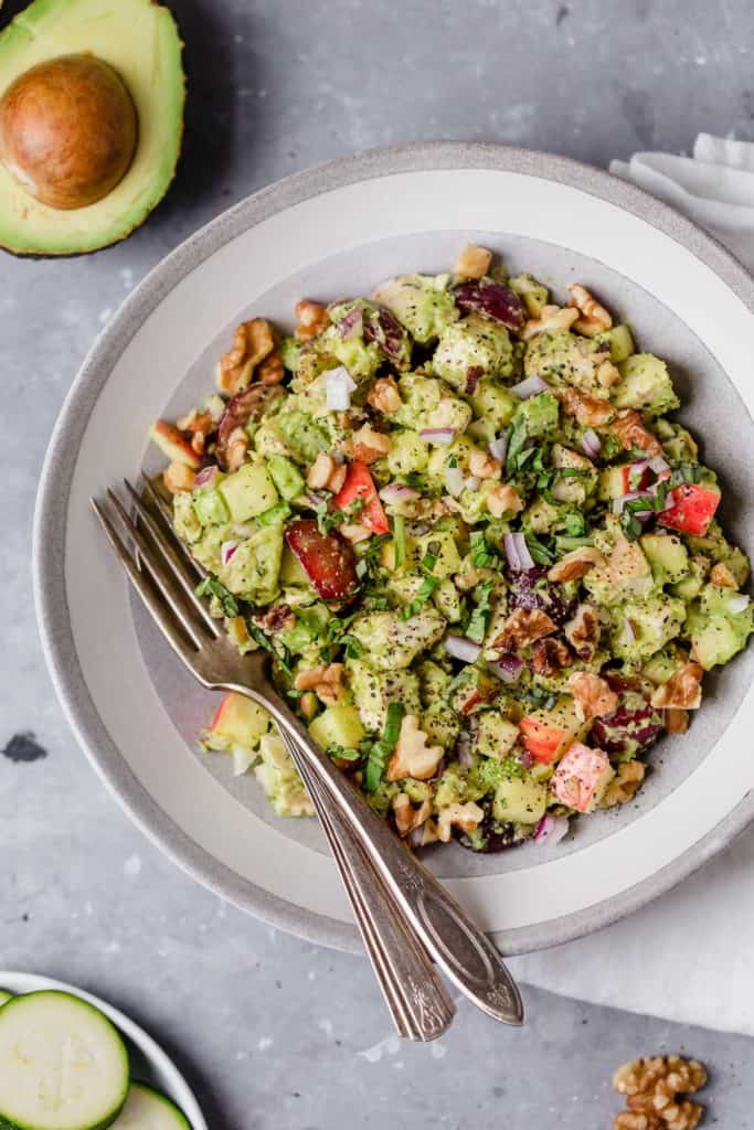 Avocado chicken salad served on plate with utensils resting in it with walnut garnish. Half an avocado as an accent.