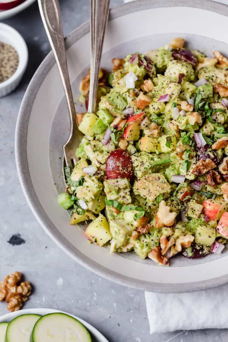 Close up view of a bowl of a chicken salad with avocado, red grapes, walnuts, and onions.