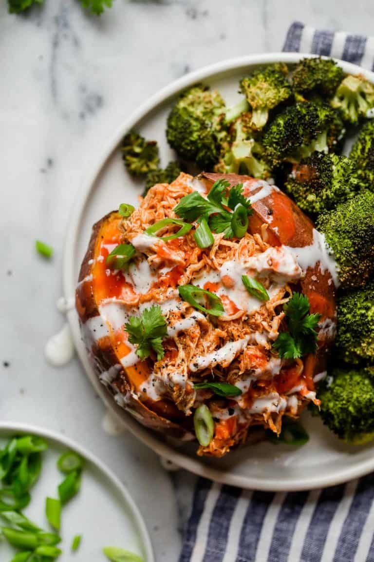Close up view of a baked sliced sweet potato filled with shredded buffalo chicken drizzled with blue cheese dressing and green onions.