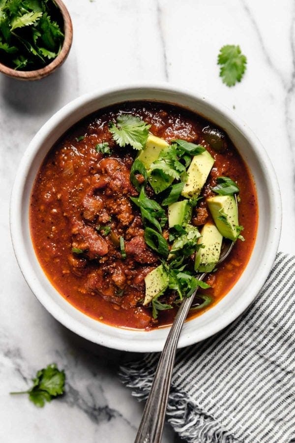 A white bowl filled with Slow Cooker Pumpkin Chili that's topped with diced avocado, fresh cilantro, and sliced green onions and a spoon in the bowl ready to eat.