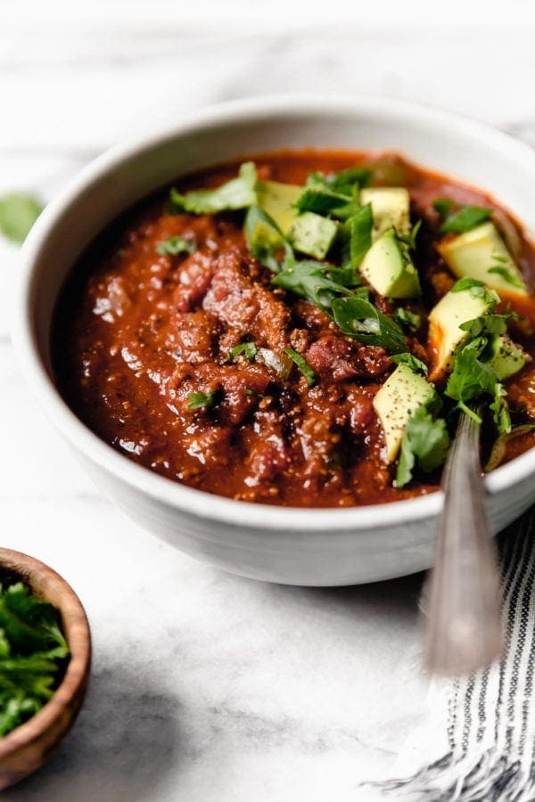 Close up/side angle view of a white bowl filled with Slow Cooker Pumpkin Chili that's topped with diced avocado, fresh cilantro, and sliced green onions. There is a spoon in the bowl so it's obviously time to eat!