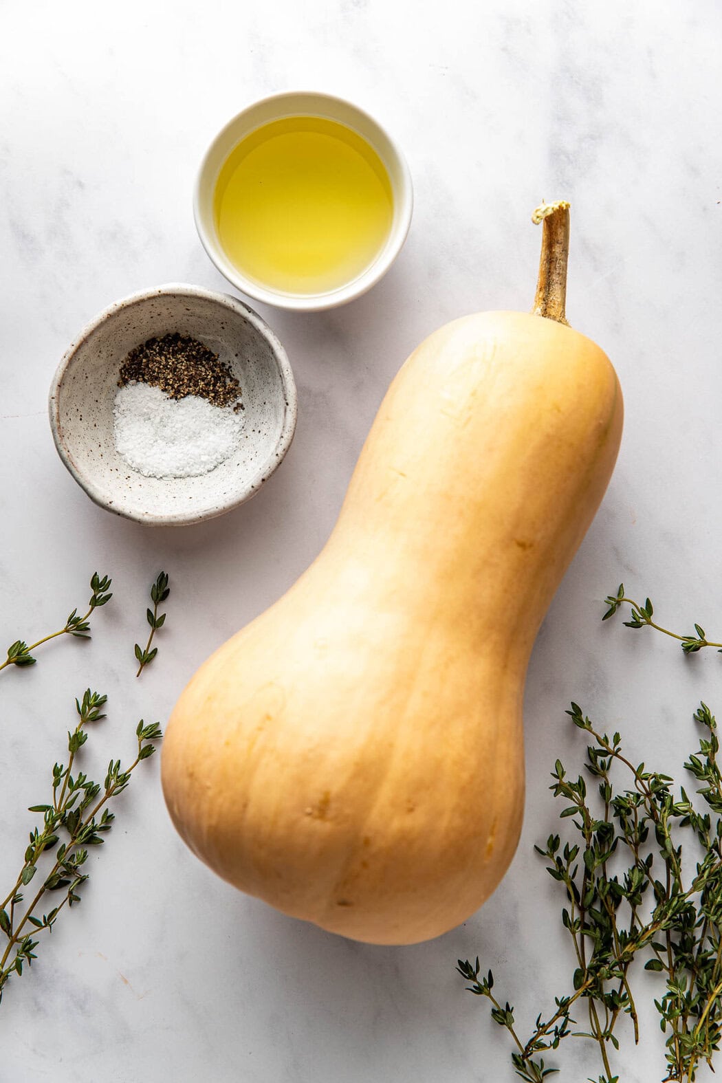 Overhead view of a white marble countertop with a butternut squash lying flat next to salt and pepper and olive oil with fresh thyme on the side.