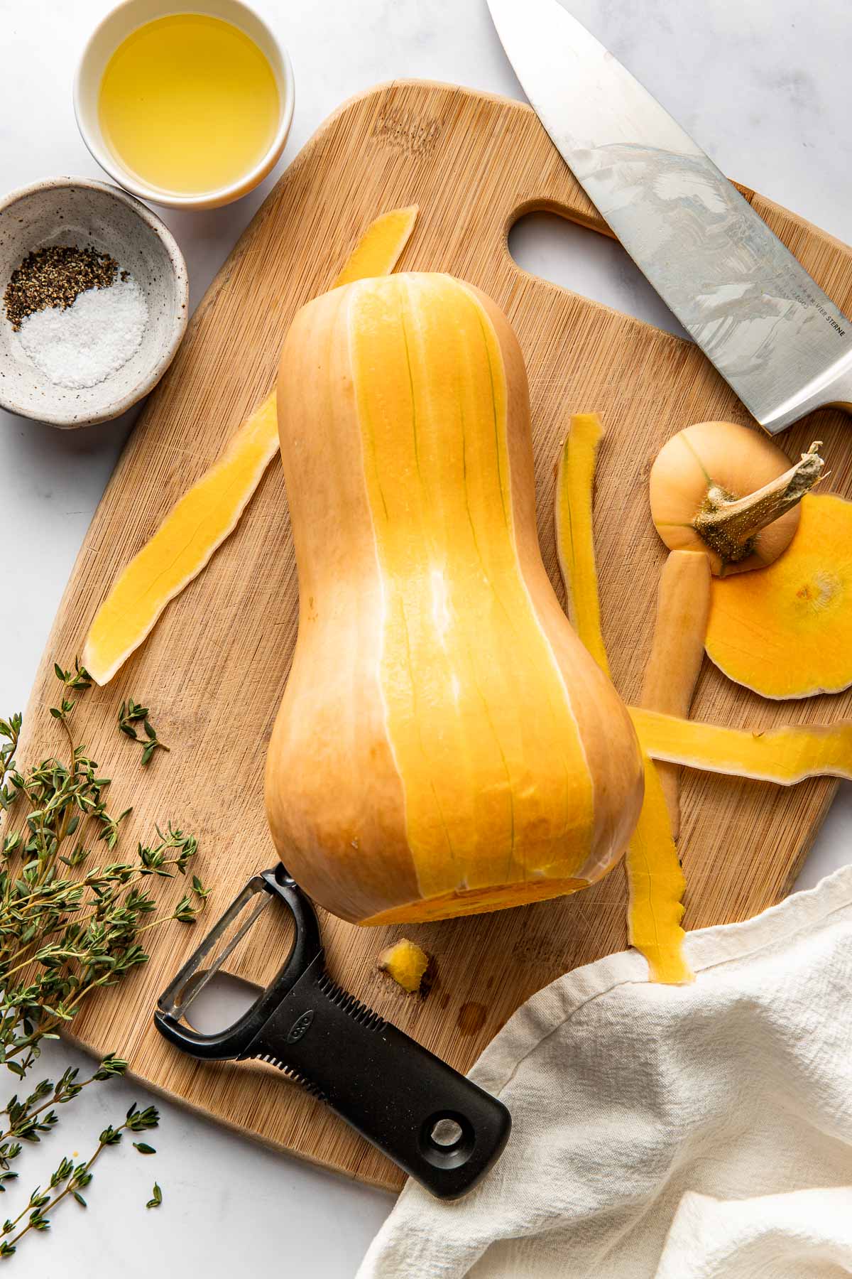 Overhead view of a wooden cutting board with a butternut squash on top cut and half peeled next to a knife. 