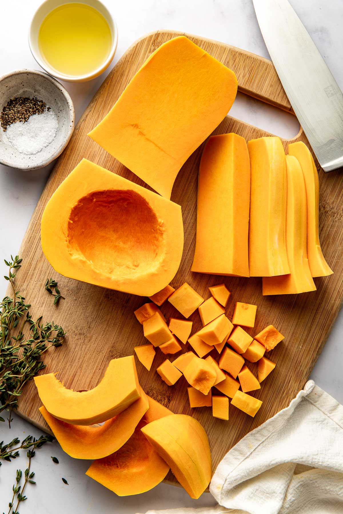 Overhead view of a wooden cutting board with a butternut squash on top that is peeled and cut next to seasonings and oil on the side. 