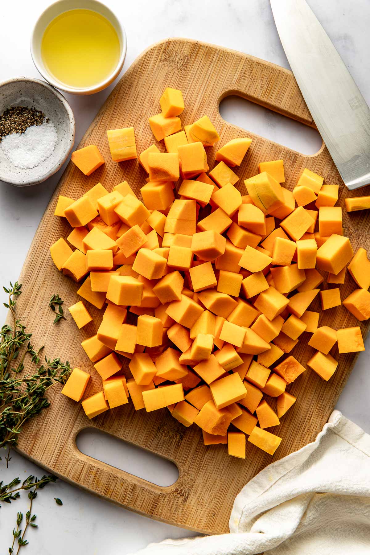 Close up view of a wooden cutting board filled with cubed pieces of butternut squash with salt and herbs on the side. 