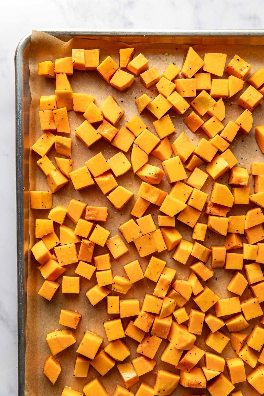 Overhead view of a sheet pan lined with parchment paper and filled with cubed butternut squash seasoned with salt and pepper. 