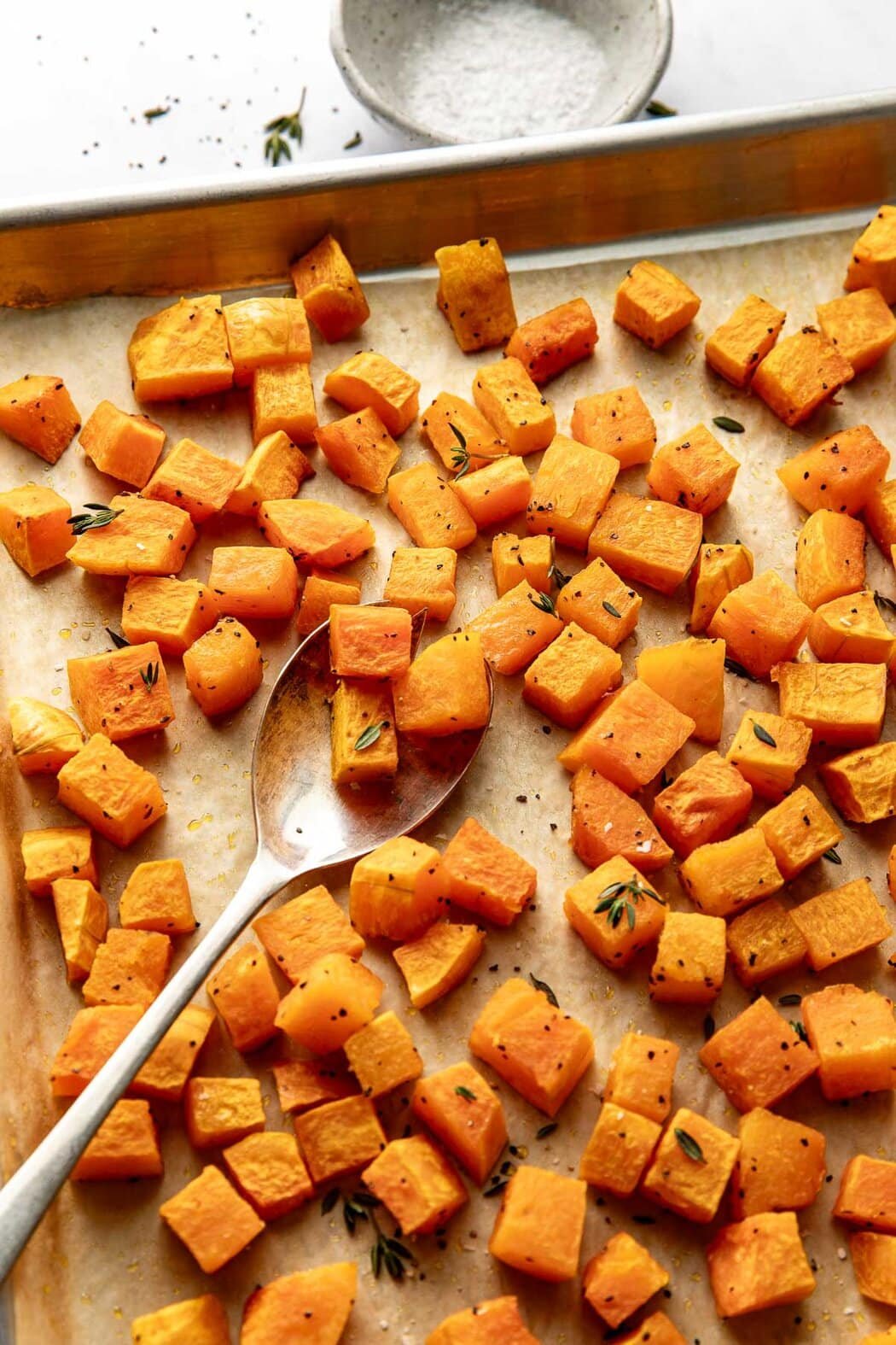 Close up view of a sheet pan lined with parchment paper filled with freshly Roasted Butternut Squash pieces topped with fresh herbs and black pepper. 
