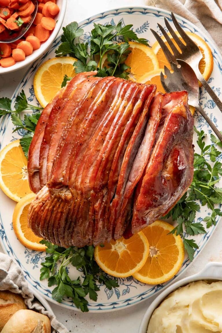 Overhead view of a large white and blue platter with a spiral cut baked glazed ham garnished with fresh greens and orange slices.