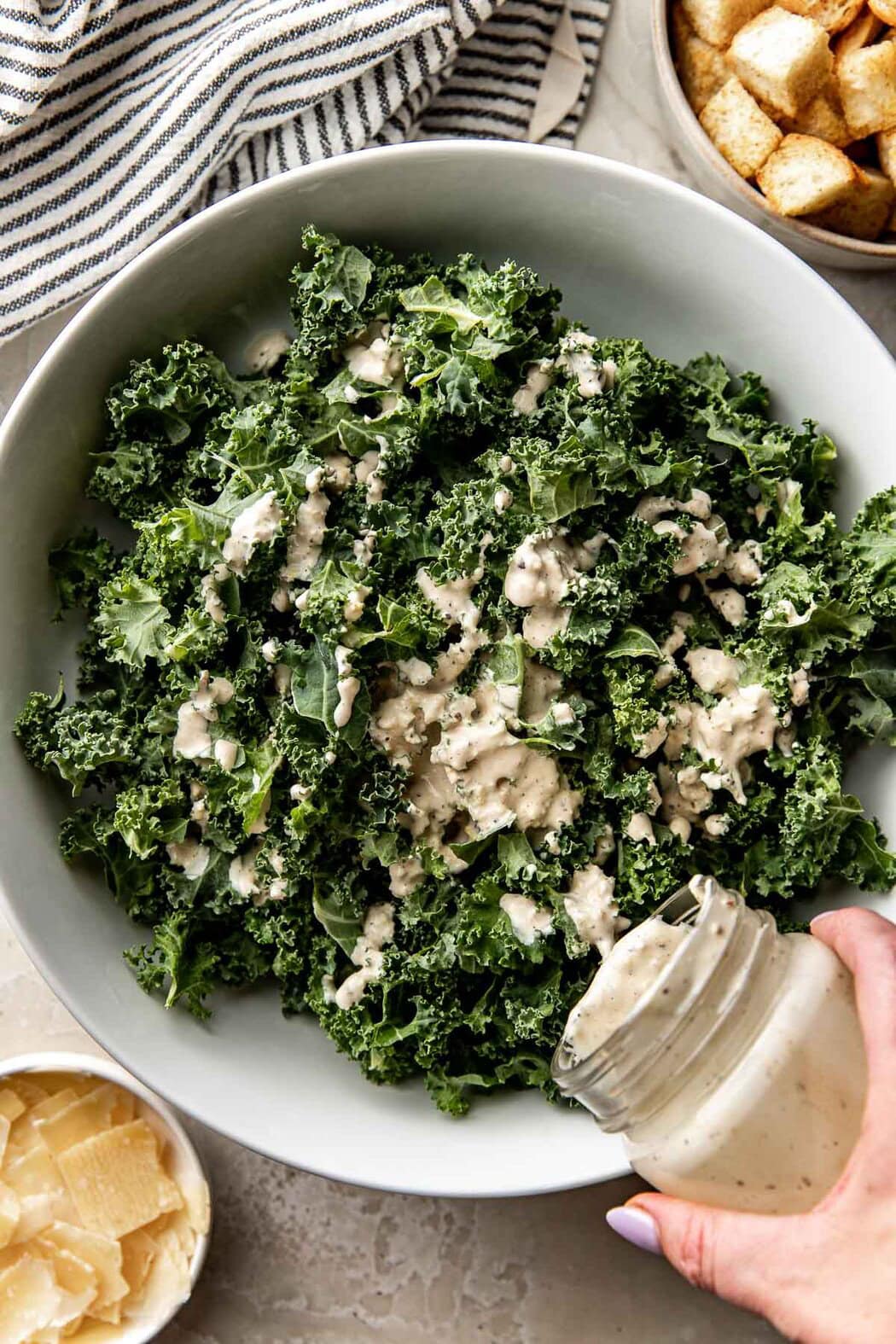 Overhead view of a hand pouring caesar dressing over a bowl of kale.