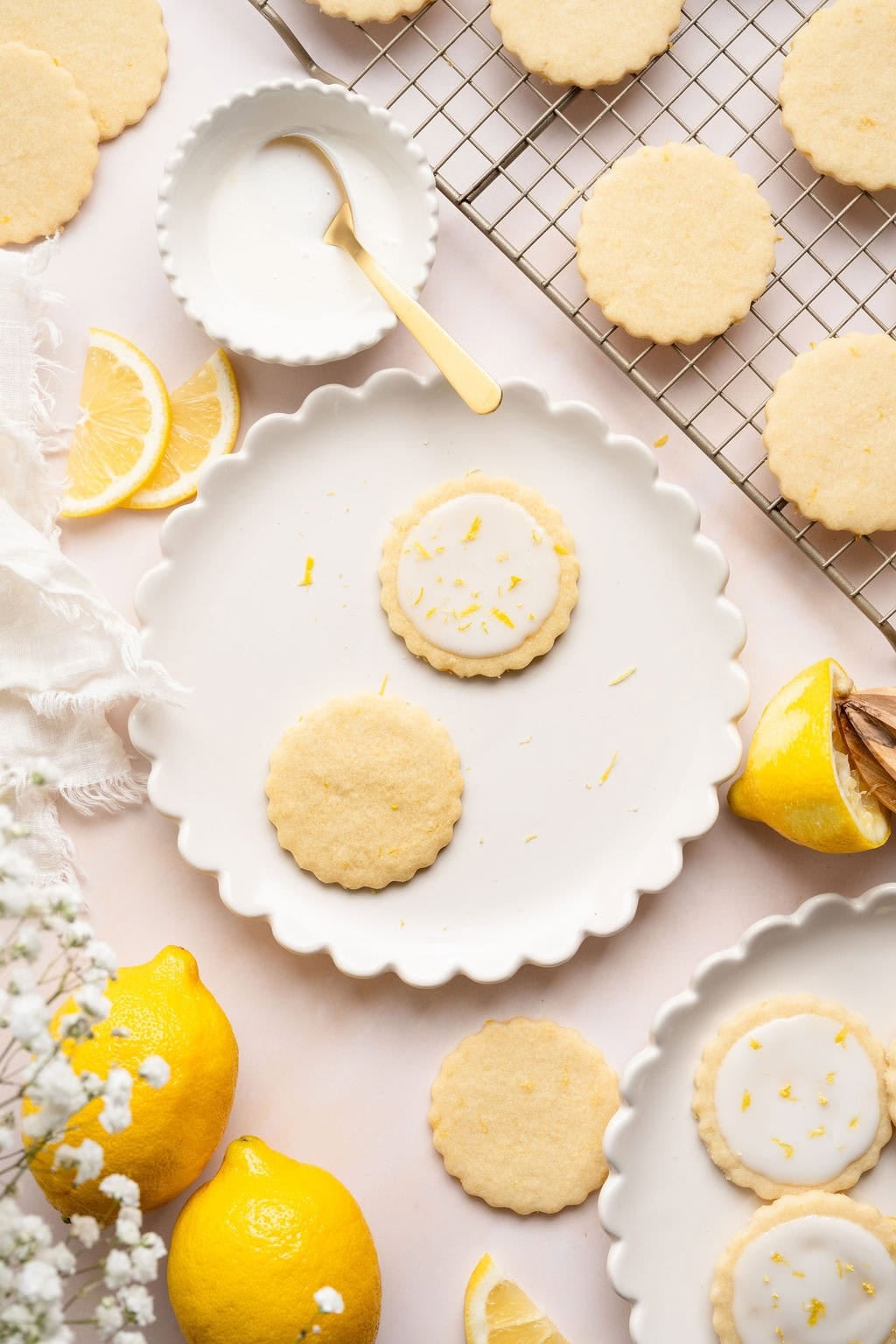 Overhead view of a lemon cookies on a serving platter next to a cooling rack with lemons arranged around the platter. 