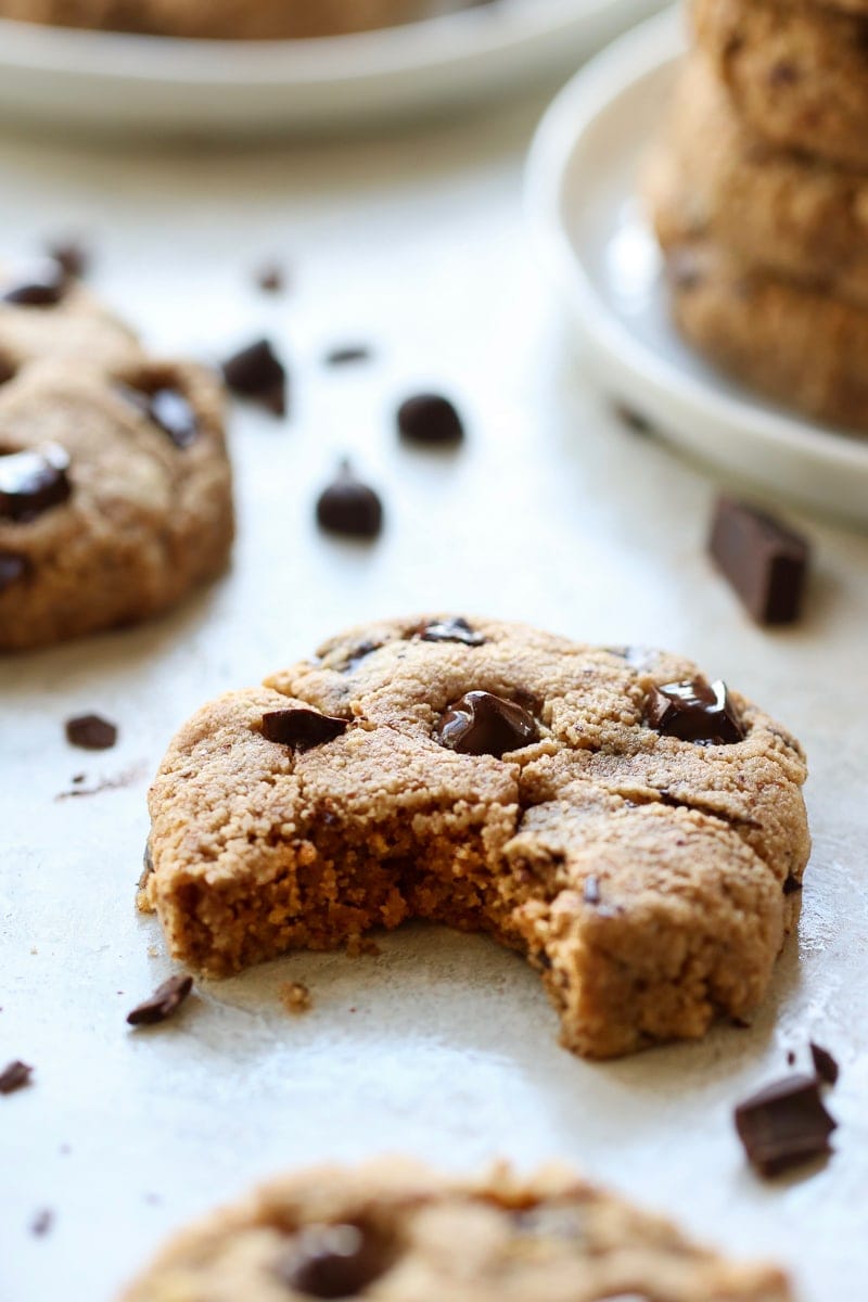 Paleo Chocolate Chip Cookies on a white platter. One of the cookies has a perfect bite out of it. Chocolate chunks and chips scattered about.