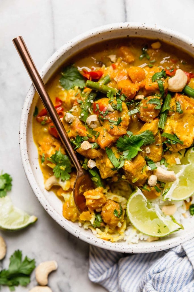 Instant Pot Chicken Sweet Potato Curry served over white rice in a white speckled bowl garnished with cilantro, lime wedges and cashews.
