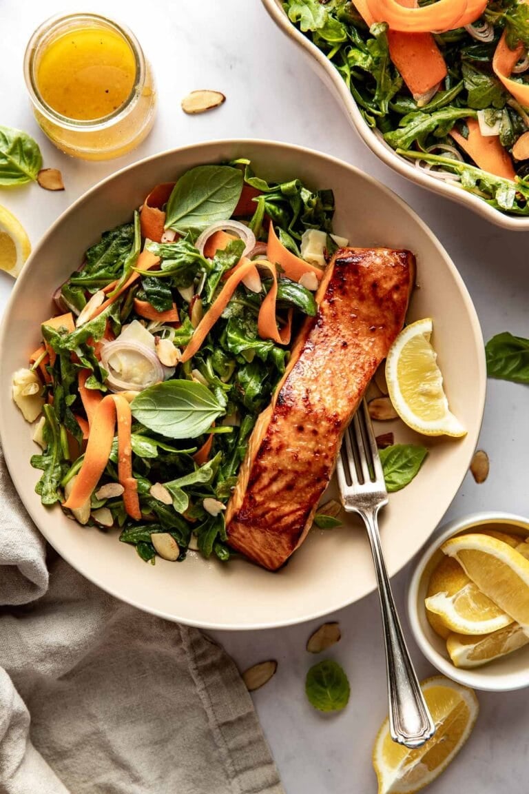 Overhead view of a bowl of arugula salad topped with fresh basil and a piece of miso-glazed salmon on the side.