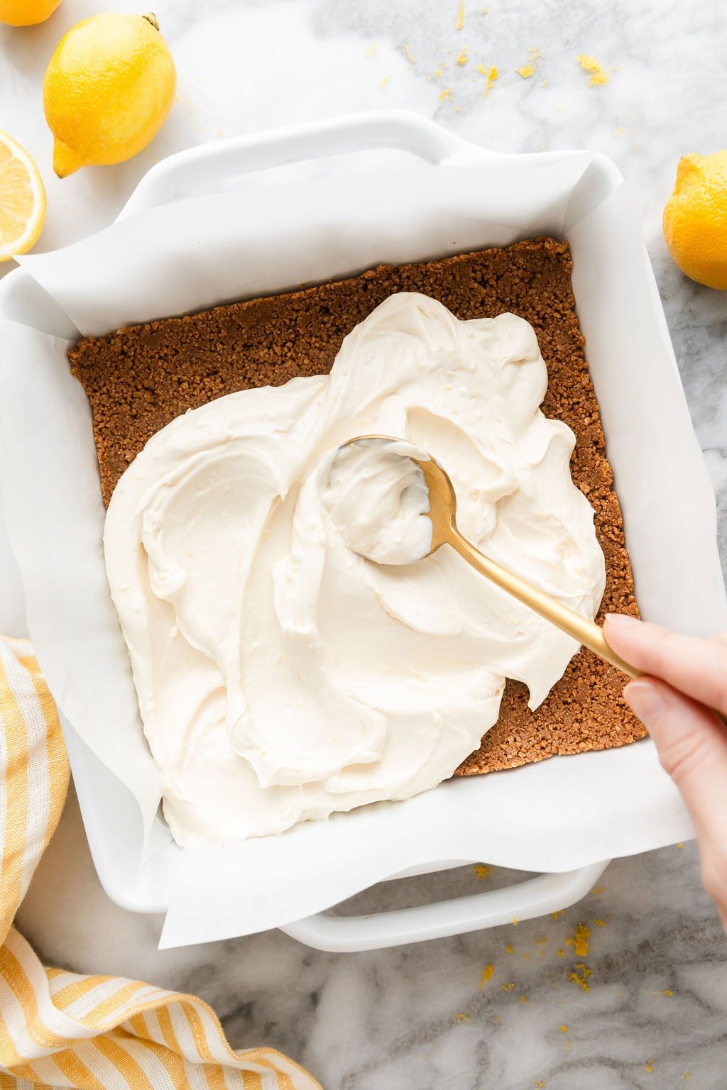 Overhead view of a pan with a hand using a spoon to spread cheesecake mixture over a graham cracker crust. 