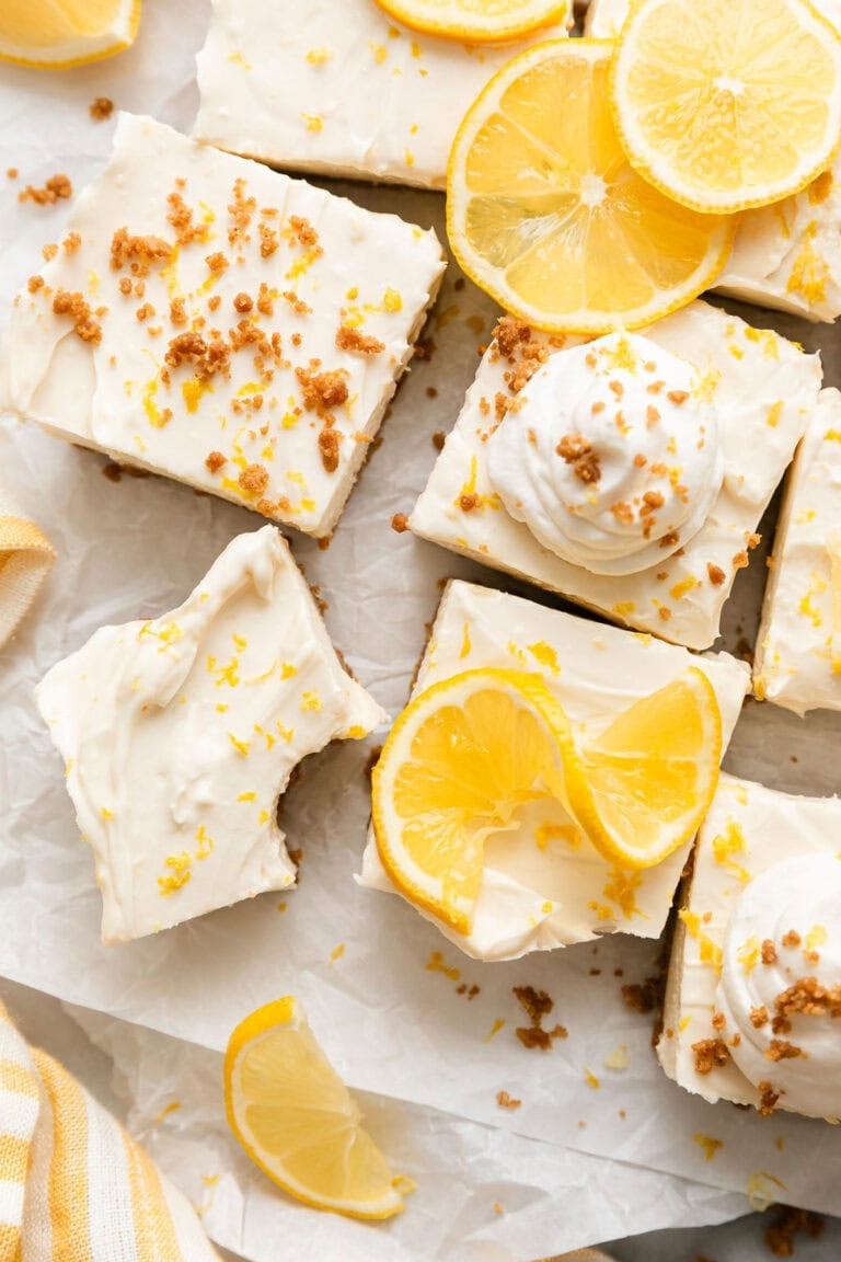 Overhead view of lemon cheesecake bars arranged on parchment paper sprinkled with a graham cracker crumble and garnished with lemons.