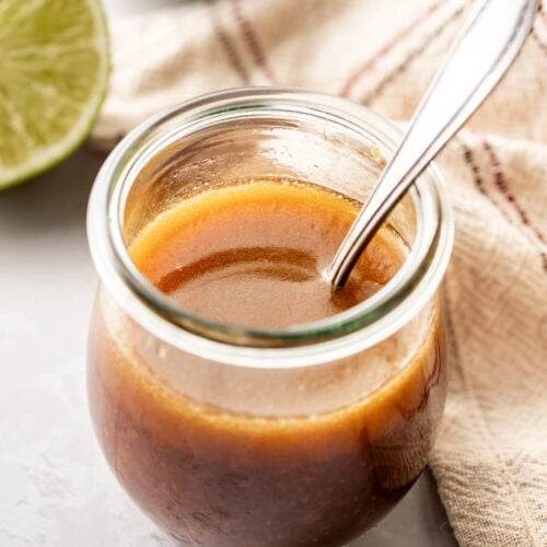 Close up view of a glass jar with a spoon in it filled with a Sesame Ginger Dressing With Lime in the background.