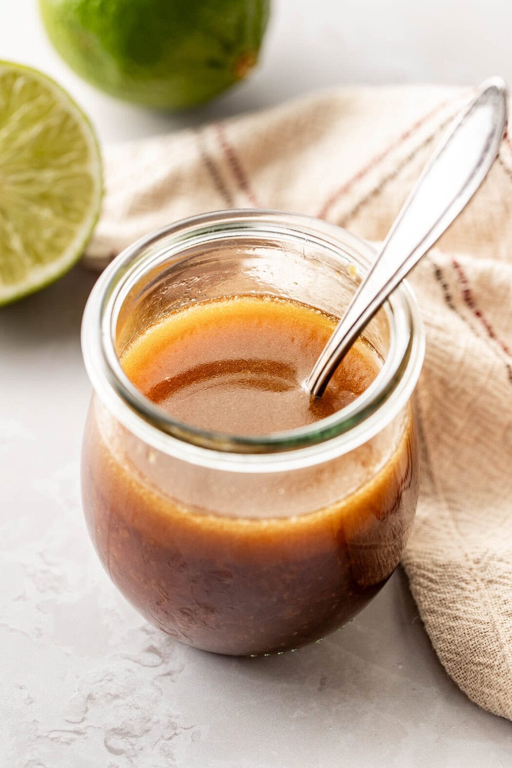 Close up view of a glass jar with a spoon in it filled with Sesame Ginger Dressing with Lime in the background. 