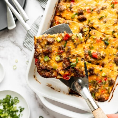 A serving spatula lifting a piece of sausage egg bake from a white casserole dish