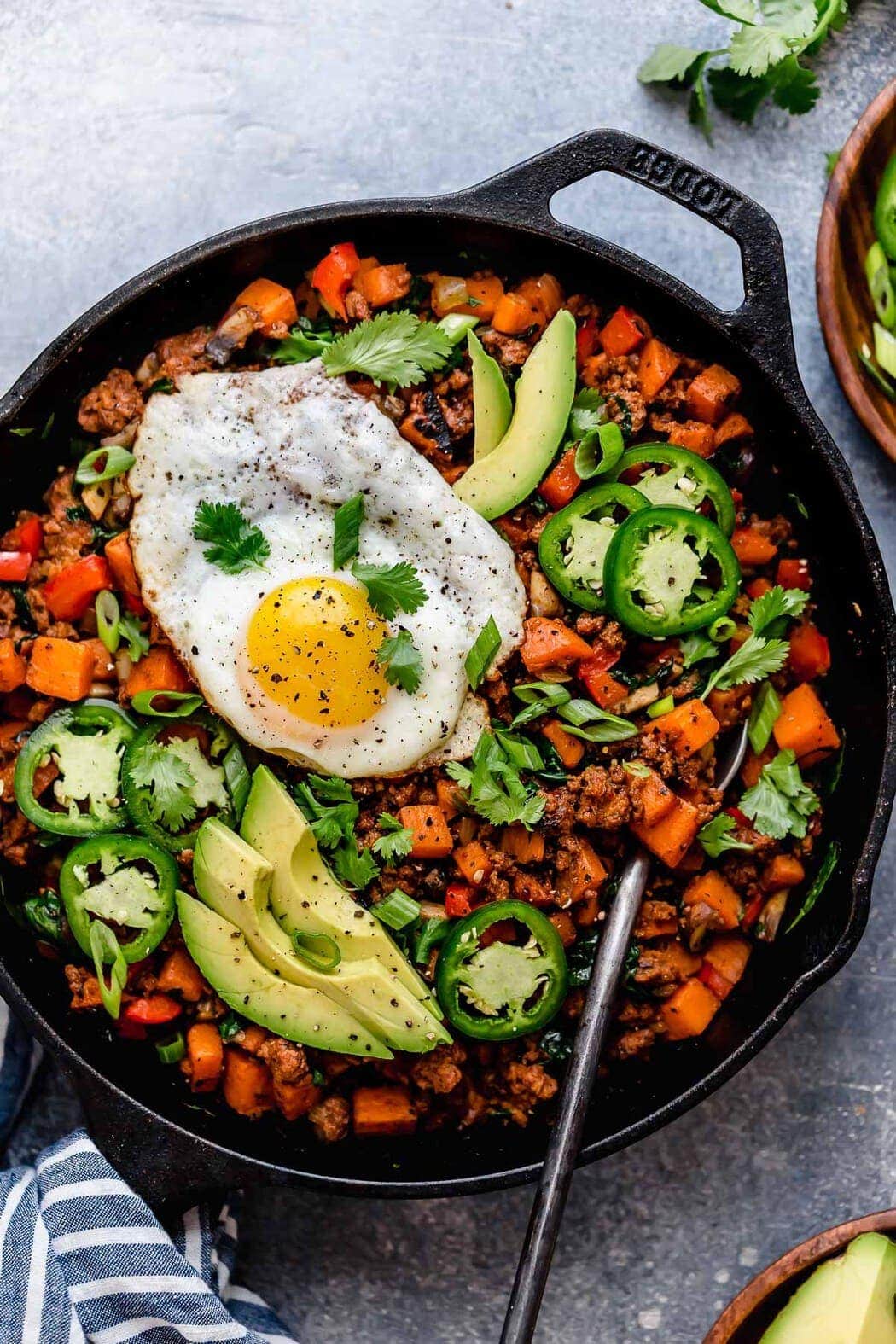 Tex-Mex Sweet Potato Hash in a black skillet with a serving spoon in it as well as a fried eggs, avocado slices, jalapeño slices, green onion and cilantro garnished over top.