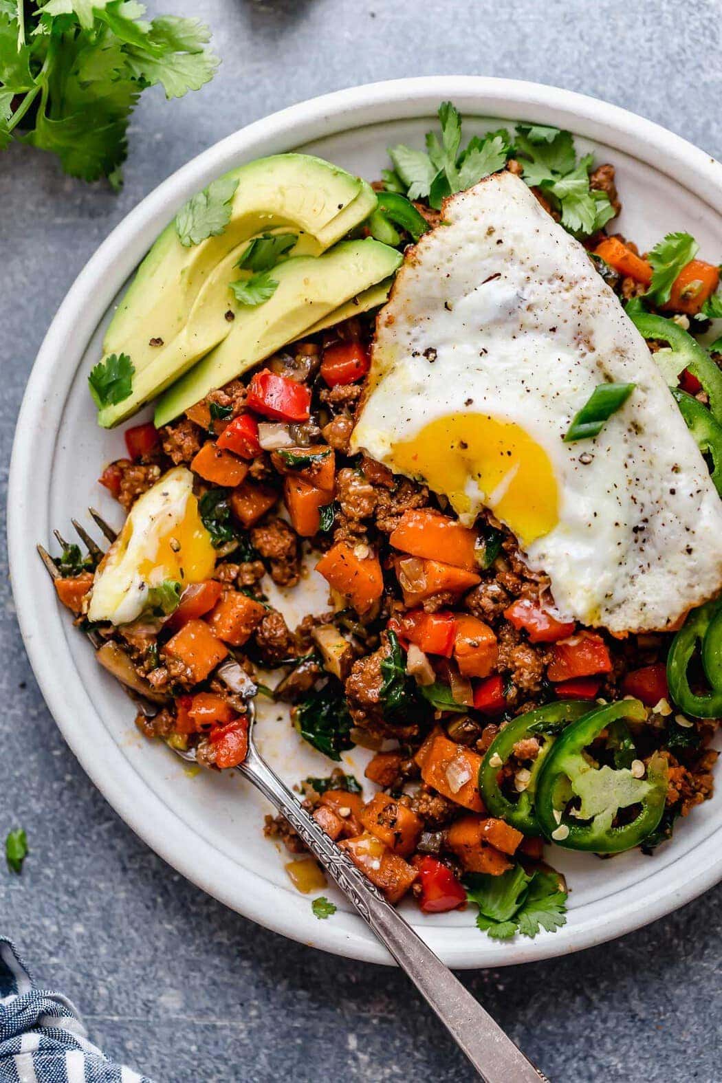 Tex-Mex Sweet Potato Hash served on a white plate with fork and a fried-egg on top with avocado slices, jalapeño slices and cilantro.
