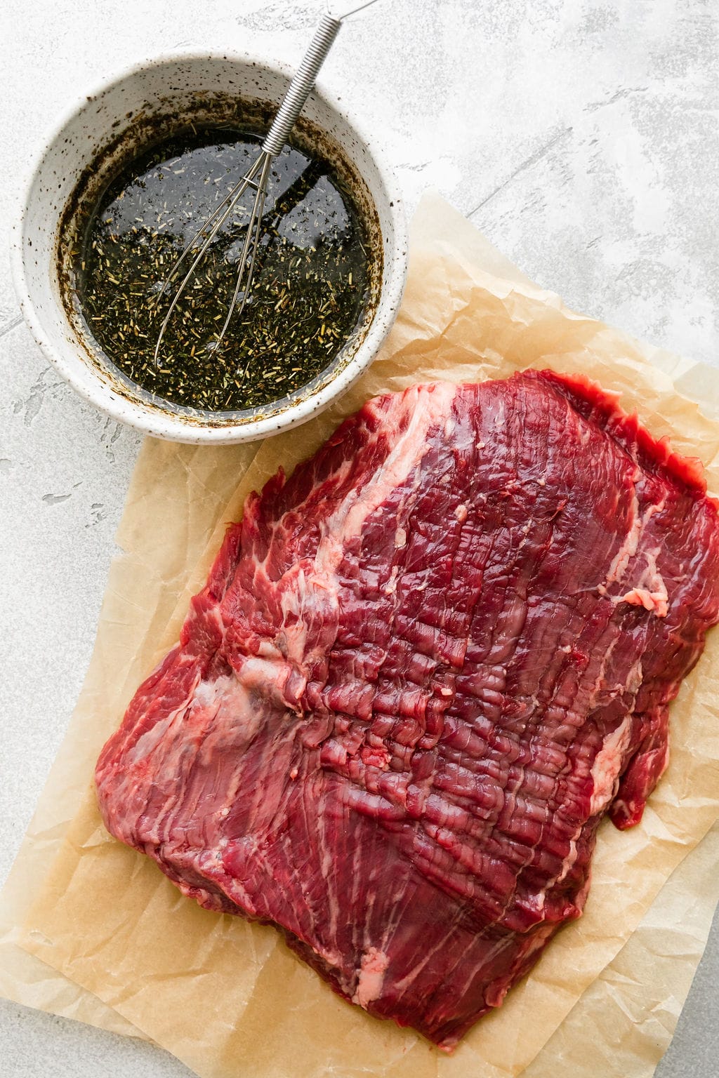 Raw flank steak on brown parchment paper with herb marinade in bowl. 
