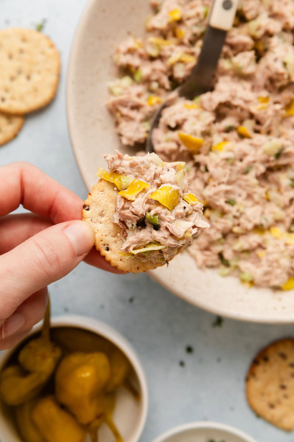 Overhead view of a hand holding a cracker topped with Tuna Salad showing pieces of chopped pepperoncini in the mixture.