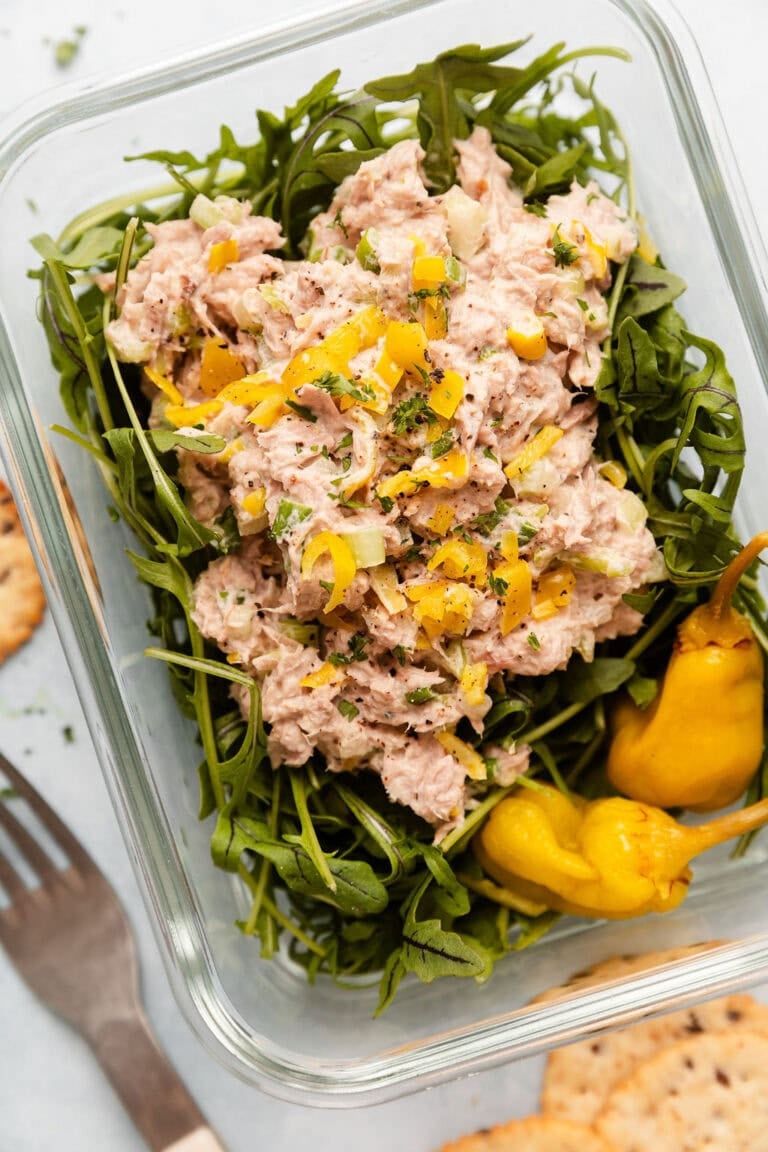 Overhead view of a glass meal prep container filled with arugula topped with tuna salad and garnished with pepperoncini peppers.