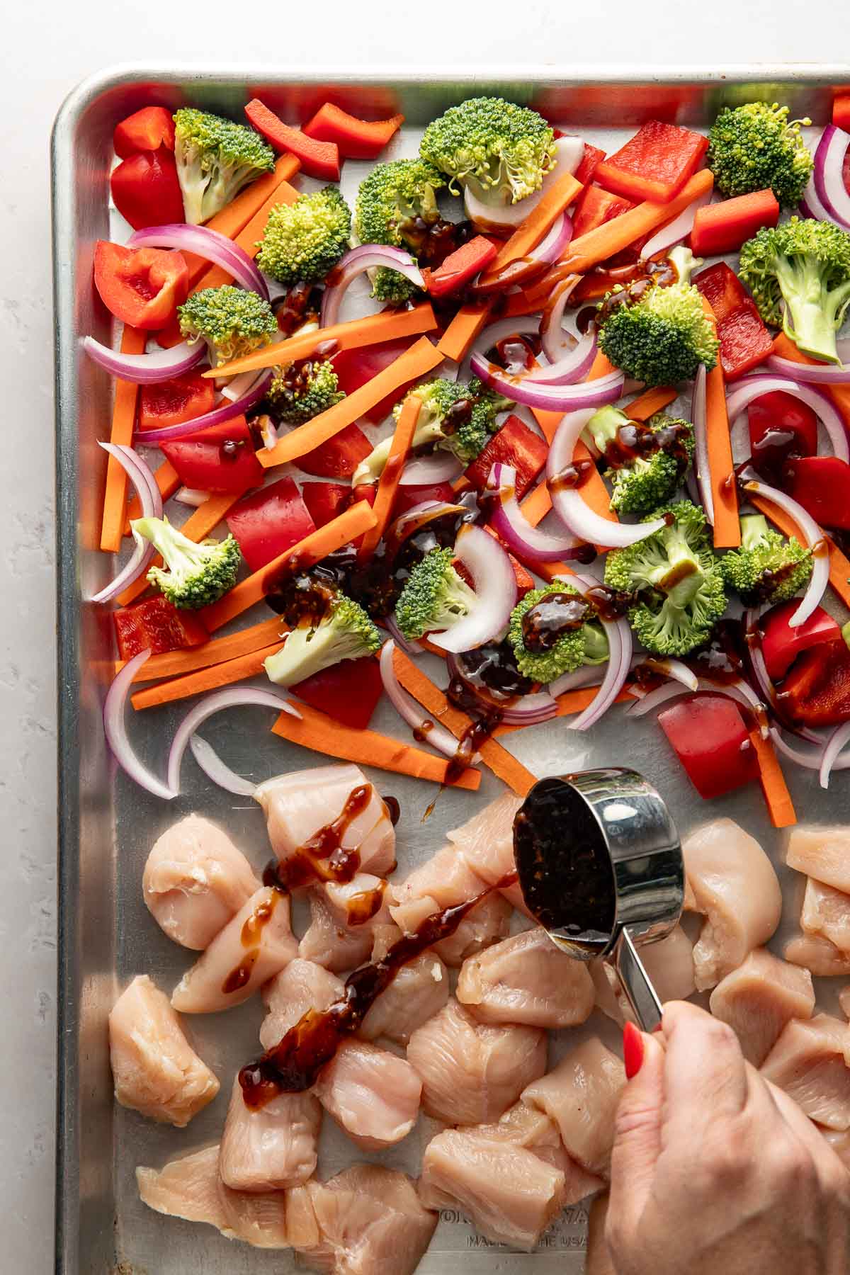 A sheet pan of raw mixed vegetables and chicken pieces, a hand drizzling teriyaki sauce on top.