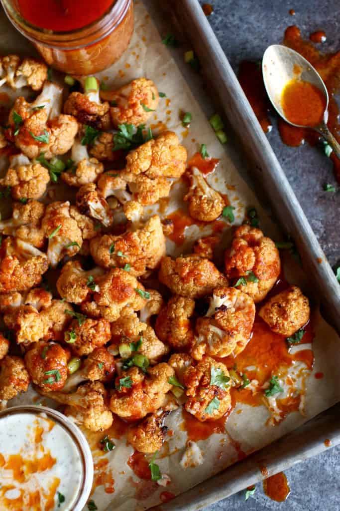 Cauliflower buffalo bites on a parchment-covered baking sheet with buffalo sauce in a jar nearby.