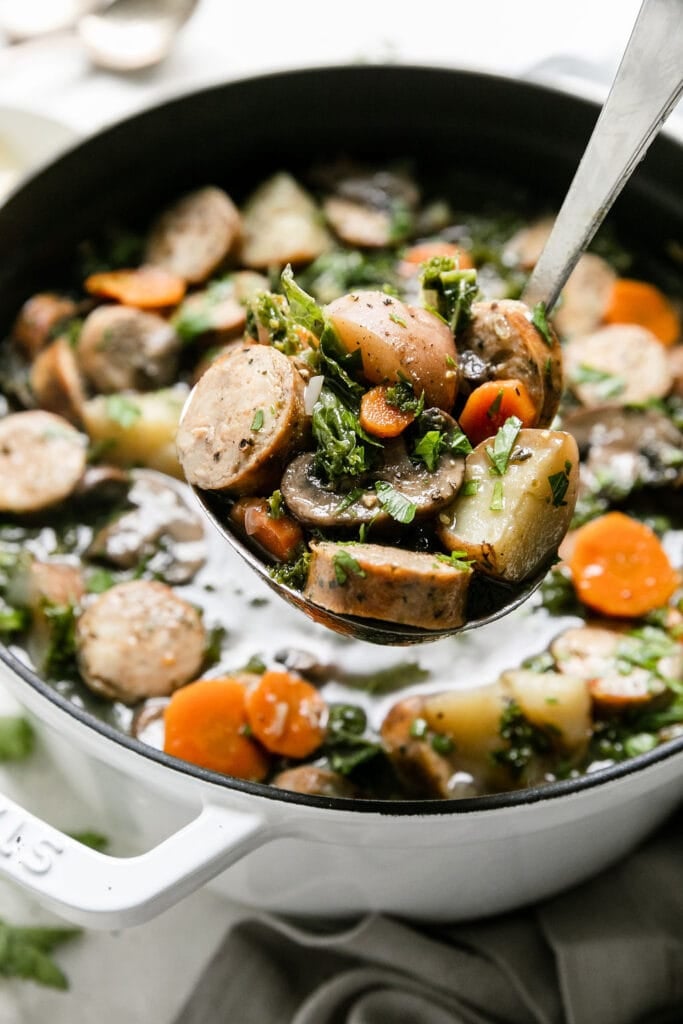 A ladle filled with Harvest Italian Soup with sausage and kale over a Dutch oven filled with soup.