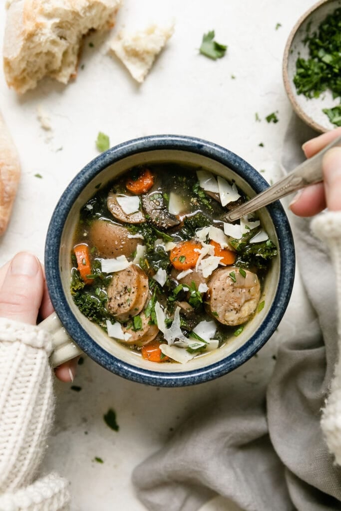 Two hands holding a blue and white bowl filled with Harvest Italian Soup with sausage, topped with shaved Parmesan cheese.