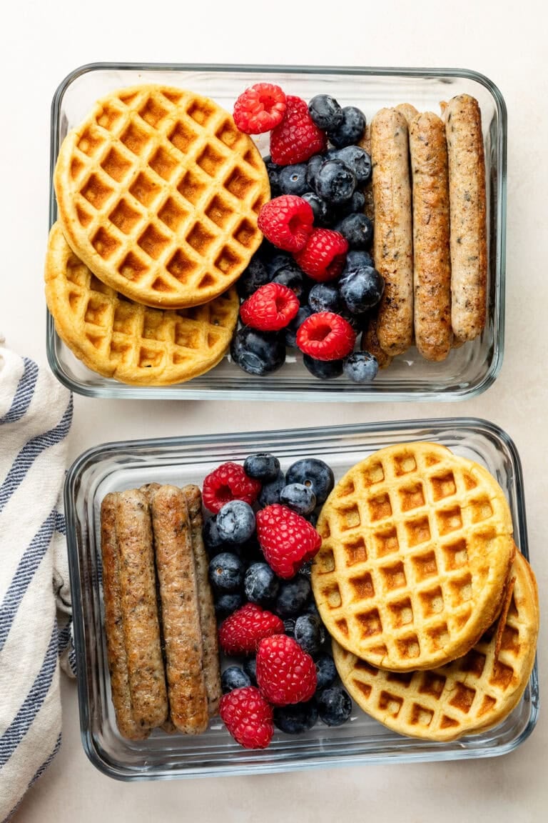 Overhead view of two glass meal prep containers filled with protein waffles, fresh berries, and sausage links.