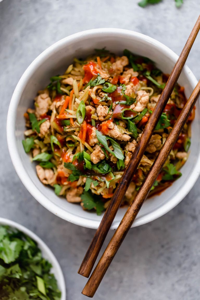 White bowl filled with broccoli slaw and ground beef asian inspired mixture topped with green onions and sriracha sauce and chopsticks for eating. 