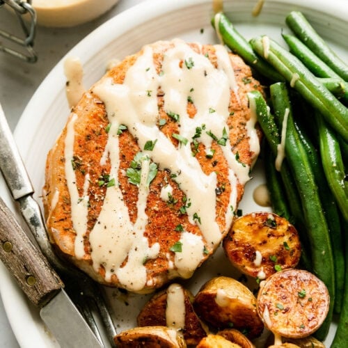 Close up view pork chop with honey mustard sauce drizzled over top, on a white plate roasted potato halves and green beans.