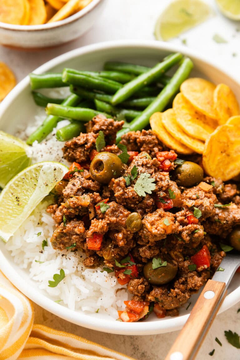 Close up view of a plate of rice topped with picadillo with a side of green beans and plantain chips topped with fresh cilantro. 