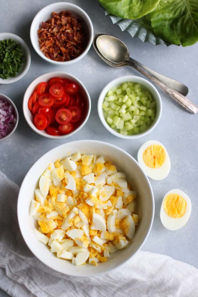 Overhead view white bowl filled with chopped hard boiled eggs, other BLT Egg Salad ingredients in individual bowls.