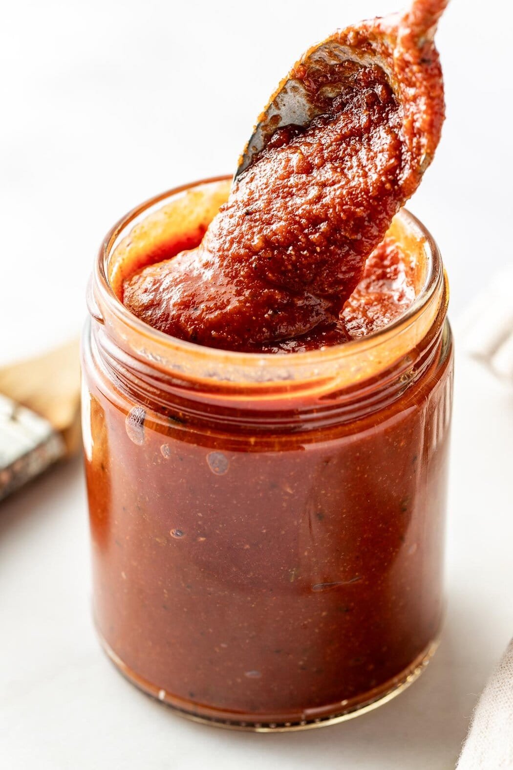 Close up view of a glass jar of Homemade BBQ Sauce with a spoon bringing out a scoop to show the thick texture.